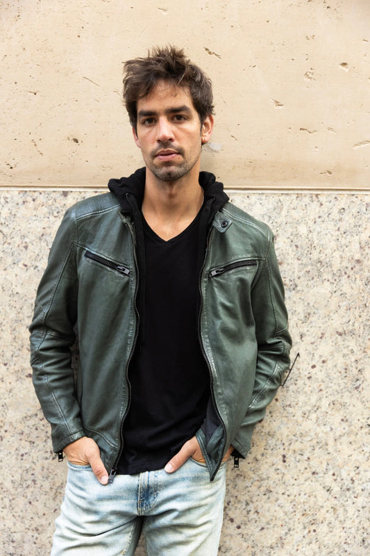 A young man with messy hair stands against a textured wall, hands in the pockets of his light-washed jeans. He is wearing a black V-neck shirt layered with the Kayto RF Leather Jacket in Dark Sage by MAURITIUS LEATHER, featuring a removable fleece hood, looking at the camera with a neutral expression.