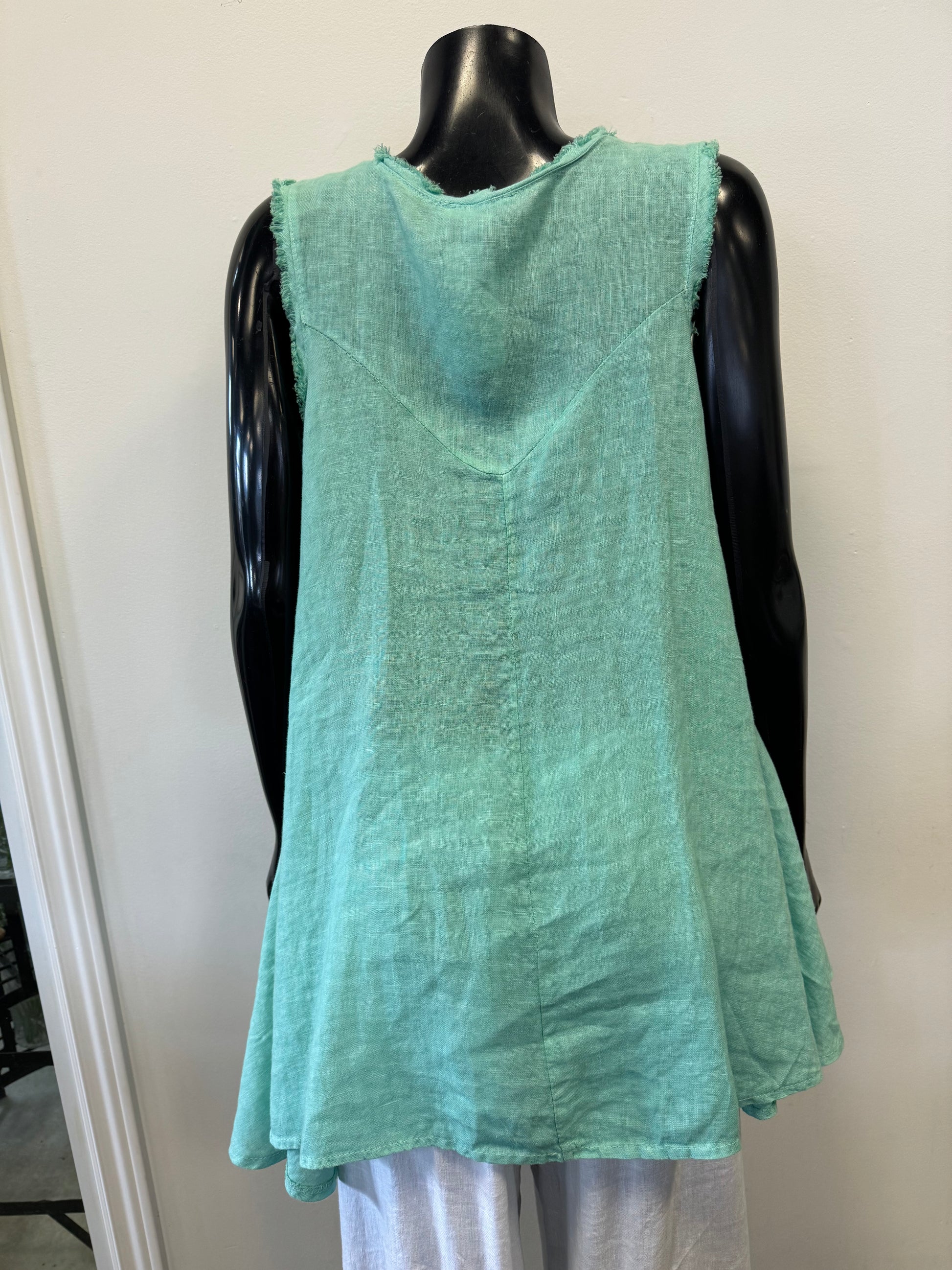 A black mannequin is wearing a Tea Green sleeveless linen top with a frayed neckline and armholes from the Catherine Lillywhite collection. The high-quality Sleeveless Linen Top - TEA Catherine Lillywhite features a loose fit that is perfect for warm weather, paired with white bottoms. The background is plain white.