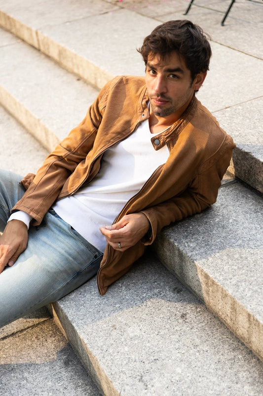 A man with short dark hair is reclining on concrete steps. He is wearing the Blake CF Leather Jacket in camel by MAURITIUS LEATHER, along with a white shirt and light blue jeans. He looks at the camera with a relaxed expression.