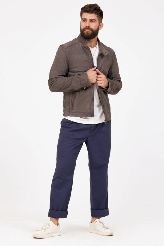 A bearded man with short hair stands against a plain white background, wearing the Bodro RF Leather Jacket in Grey by MAURITIUS LEATHER, a white t-shirt, dark blue cuffed pants, and white sneakers. His hands are adjusting the collar of his spring jacket as he looks to the side.