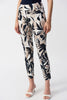 Wearing Joseph Ribkoff’s Millennium Tropical Print Slim Fit Pants 251001, which feature a high-waisted, bold black, white, and beige leaf pattern paired with a navy top and white open-toe heels. The outfit exudes elegance with one hand resting casually by their side.