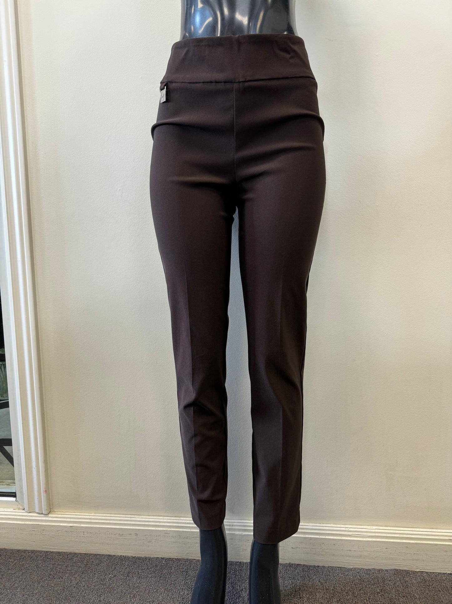 A pair of Joseph Ribkoff Pants Style 201483TT MOCHA in dark brown is displayed on a black mannequin against a plain white wall. These dressy yet comfortable trousers feature a high waist and front seam detailing, with a small pocket visible on the right side. The mannequin stands on a grey carpeted floor.