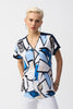 A short-haired person with blonde hair models the Joseph Ribkoff Silky Knit Geometric Print Straight Top 251246, featuring blue and white patterns and a front zipper, paired with white pants. They stand against a plain background, showcasing a neutral expression.