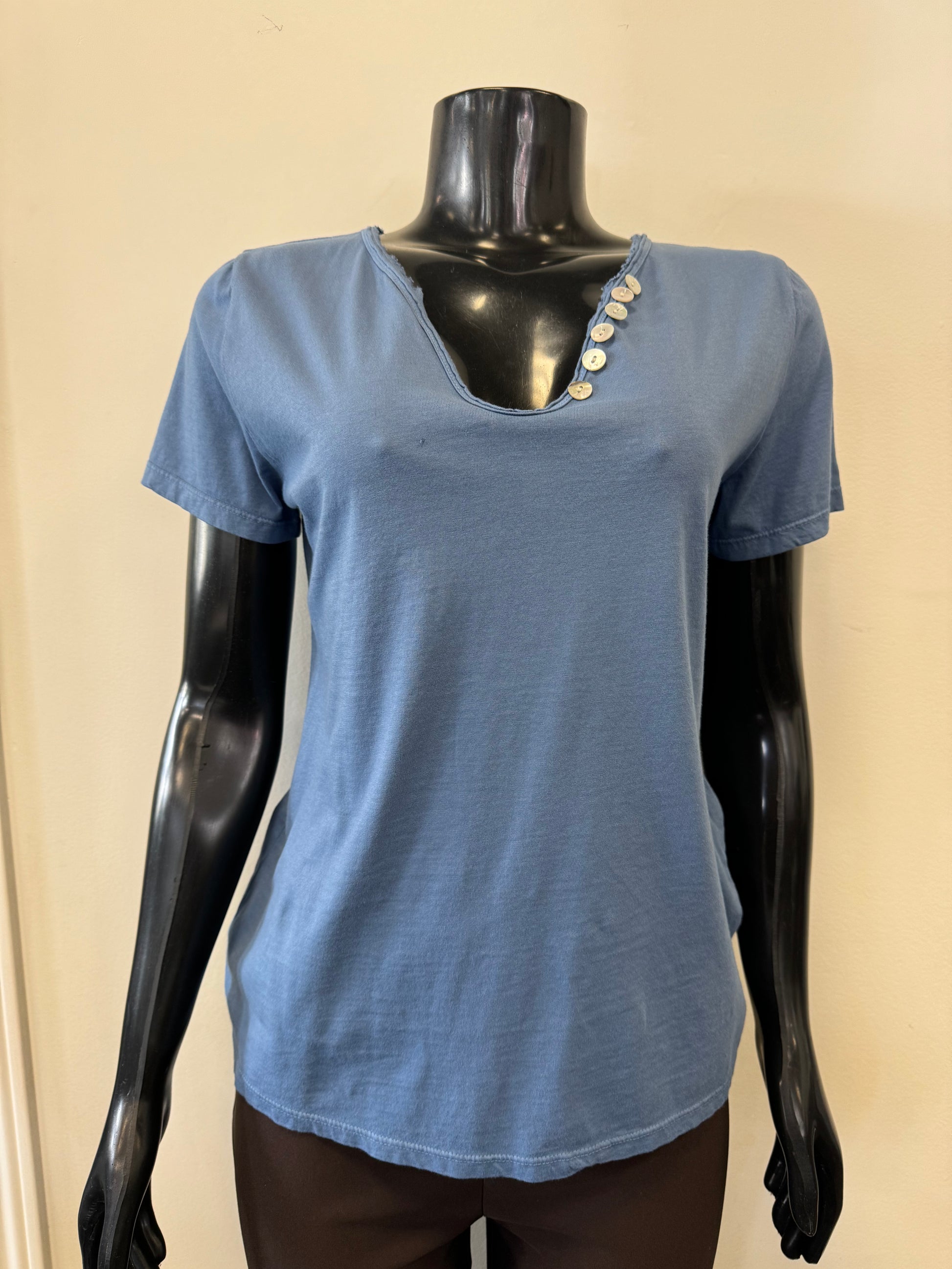 The elegant Femme Fatale Indigo Cotton Top is showcased on a black mannequin. This Italian-made shirt features short sleeves, a notched neckline, and four decorative buttons on one side. It hangs loosely with a slight flare at the bottom against a plain off-white background.