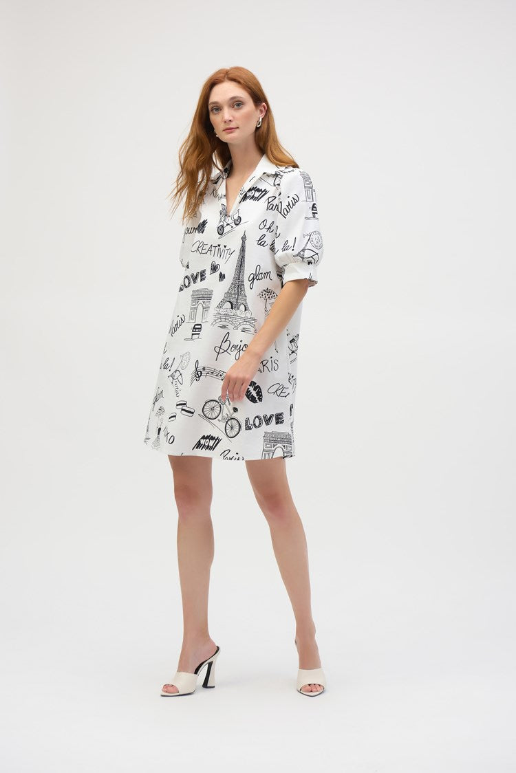 A woman with long red hair wears the Joseph Ribkoff Poplin Conversational Print A-Line Dress 252019, featuring black text and Paris-themed illustrations. She pairs it with white heels while posing gracefully against a plain white background.