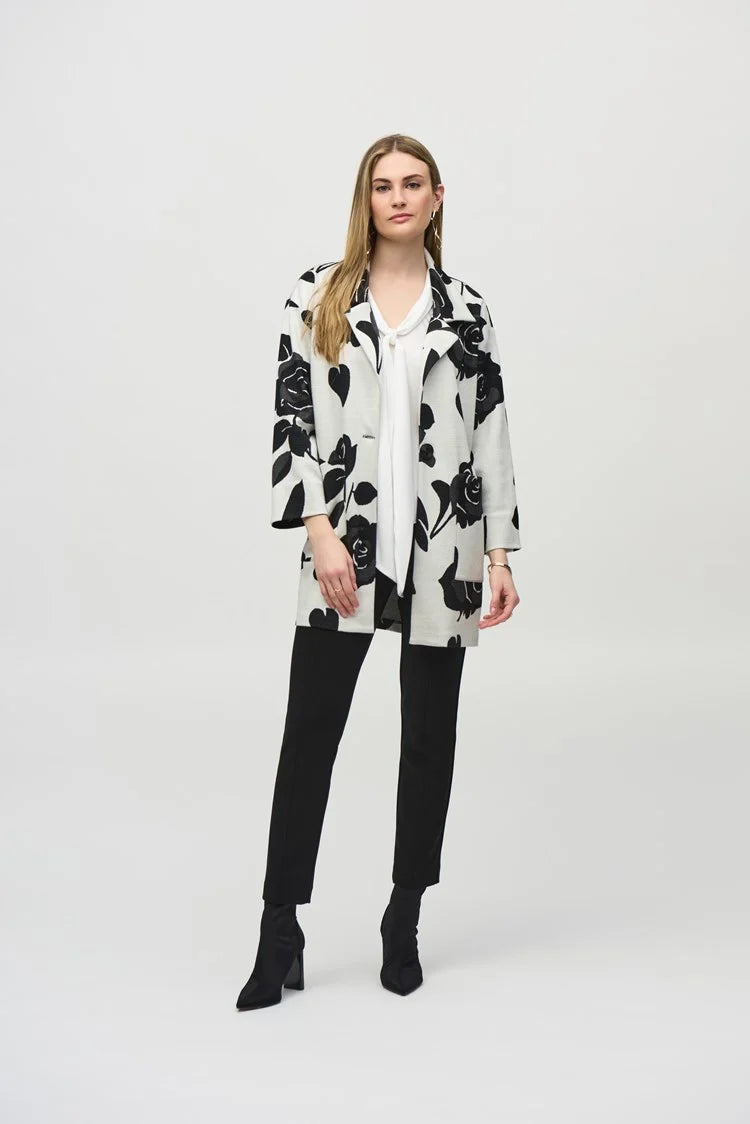 A person wearing the Jacquard Sweater Floral Straight Coat 244099 by Joseph Ribkoff, featuring a white floral pattern and a notched collar, is styled over a white blouse and paired with black trousers and ankle boots, standing against a plain background.