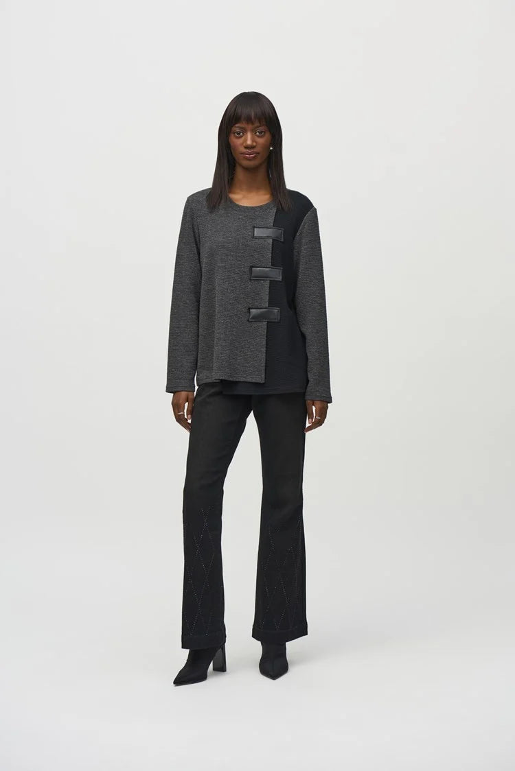 A person stands against a plain background wearing the fashionable Textured Knit Color Block Boxy Tunic by Joseph Ribkoff, characterized by its asymmetrical gray design with black accents and distinctive fastening details, including a leatherette patch. The textured knit offers depth and is expertly paired with black pants and pointed black shoes.