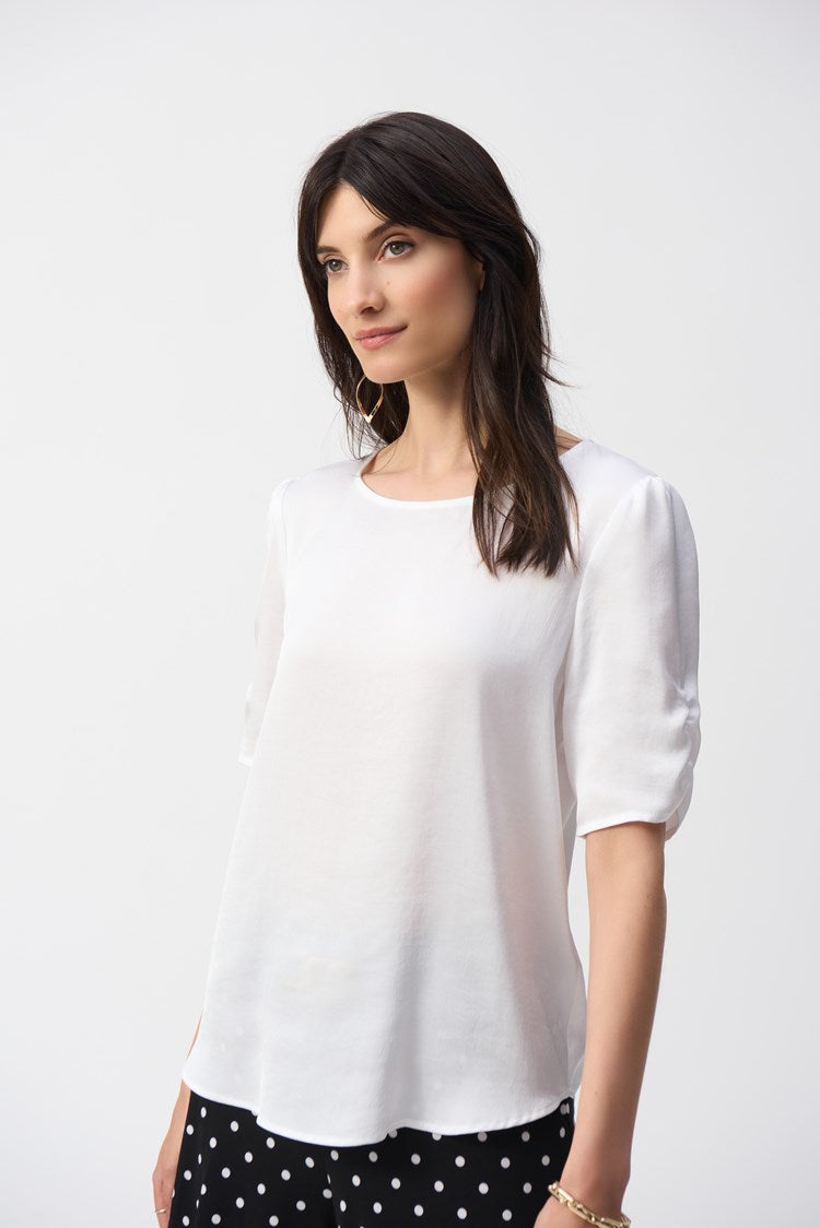 A woman with long dark hair wears the Joseph Ribkoff Hammered Satin Straight Top 251119, featuring shirred short sleeves, paired with black polka dot pants. She stands against a plain white background, looking to the side with a neutral expression.