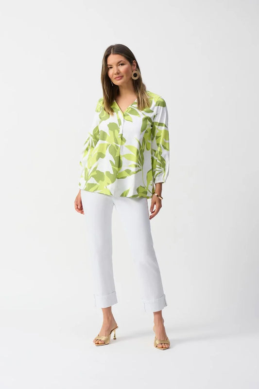 A woman stands against a plain backdrop wearing the Joseph Ribkoff Stretch Poplin Floral Print Trapeze Top 251168 with three-quarter sleeves, paired with white pants and gold heels. She has long brown hair and accessorizes with large hoop earrings.