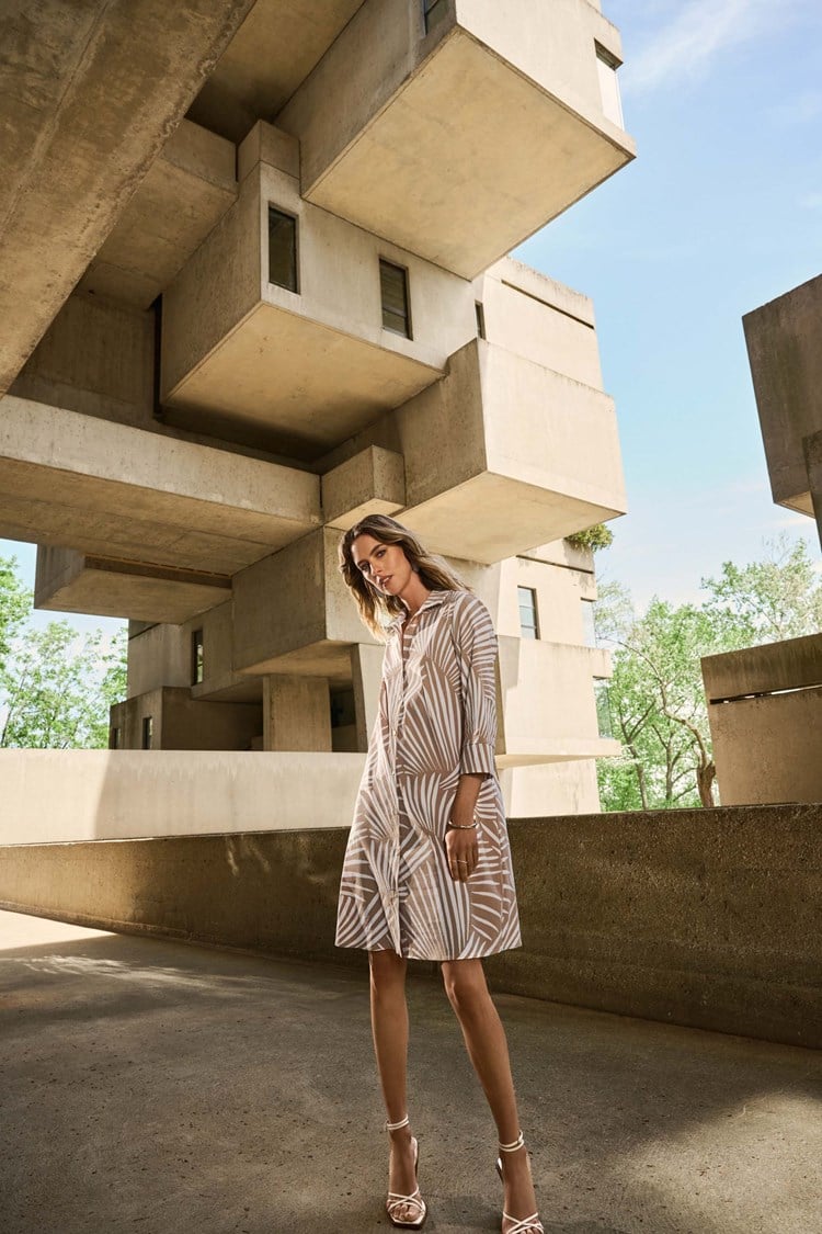 Wearing Joseph Ribkoff's Stretch Poplin Tropical Print Shirt Dress 251144 paired with white sandals, a person complements the block-like structures above. The dress sways gently, enhancing the serene backdrop of greenery and blue sky.