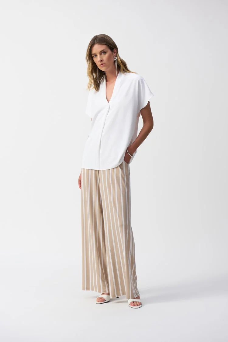 A person is styled in Joseph Ribkoff's Striped Silky Knit Wide-Leg Pants 241135S25 and a white short-sleeved blouse, posed against a plain white background. They accessorize with earrings and sandals, with one hand in their pocket.