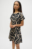 A long-haired individual wears the Seersucker Abstract Print A-Line Dress 252205 by Joseph Ribkoff, featuring short sleeves and a striking black-and-cream pattern against a plain white background, standing with hands clasped.