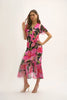 Against a light background, a woman poses in the Joseph Ribkoff Silky Knit and Chiffon Floral Print Wrap Dress, featuring vibrant pink flowers and ruffled chiffon sleeves in a flounce silhouette. She completes the look with strappy heels, one hand on the wall and the other on her hip.