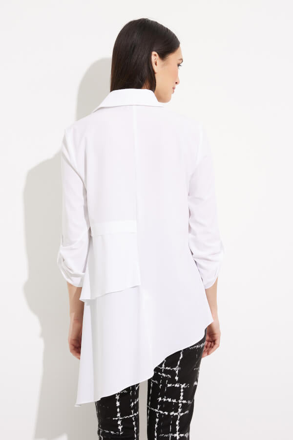 A person with long dark hair is wearing a white Joseph Ribkoff Tiered Asymmetric Hem Blouse Style 233099, which features asymmetric layers on the front and 3/4 sleeves. Paired with black pants adorned with a white grid pattern, this stylish ensemble by Joseph Ribkoff is perfect for an office wardrobe. The background is plain white.