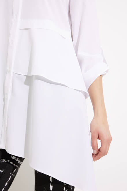 A person with long dark hair is wearing a white Joseph Ribkoff Tiered Asymmetric Hem Blouse Style 233099, which features asymmetric layers on the front and 3/4 sleeves. Paired with black pants adorned with a white grid pattern, this stylish ensemble by Joseph Ribkoff is perfect for an office wardrobe. The background is plain white.