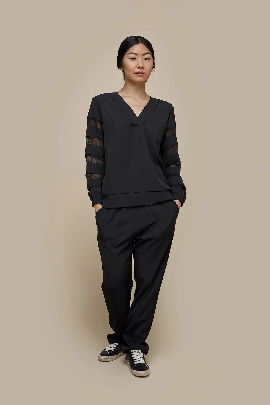 A person with dark hair is wearing the UCHUU Loose Long-Sleeved Sweater with Yoke Details (CS24-732) in black, featuring sheer long sleeves with horizontal stripes. They are also wearing black pants and black and white sneakers, standing against a plain beige background.