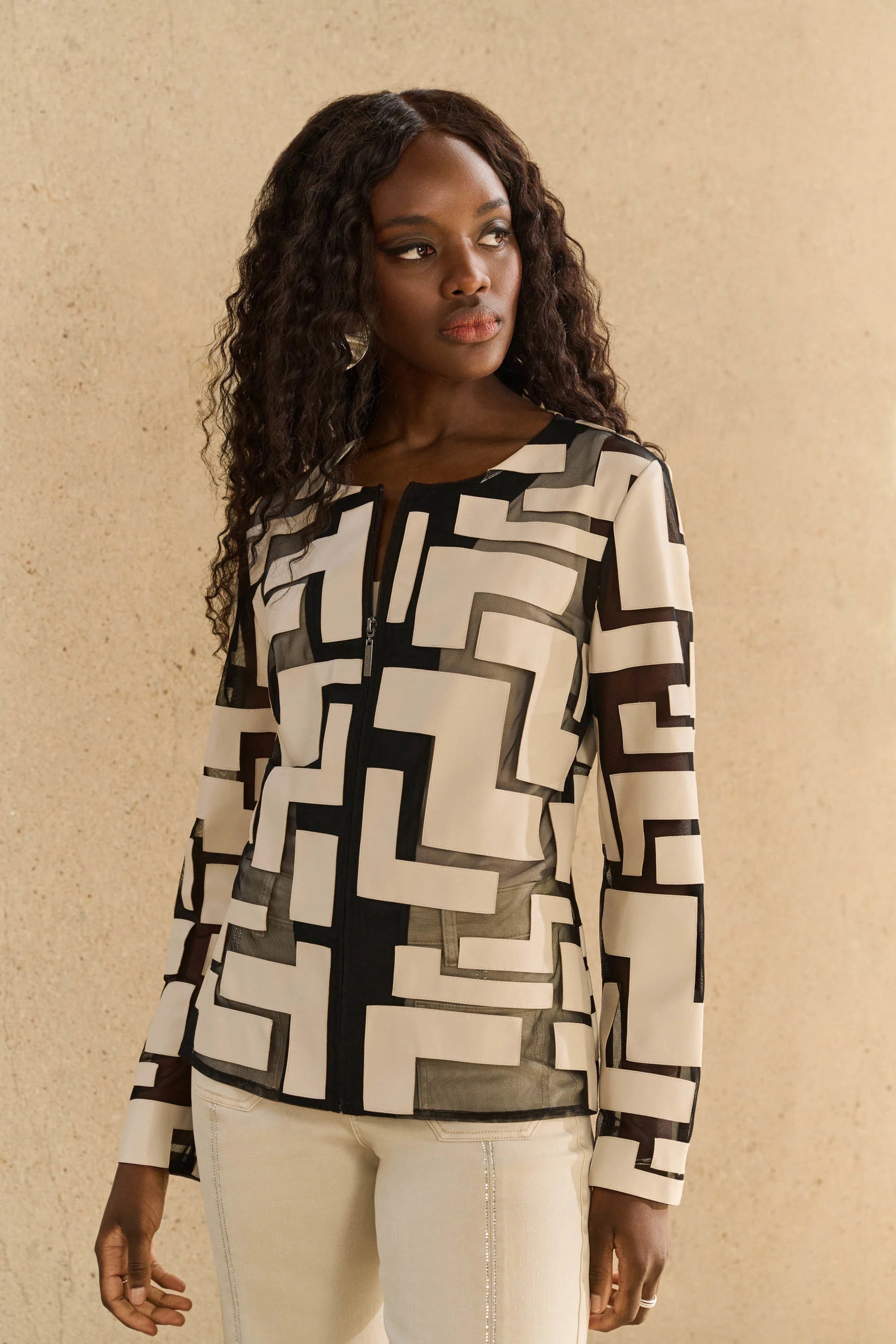 A woman with long curly hair wearing a Joseph Ribkoff geometric-patterned black and beige top with a crew neckline stands against a light brown wall, looking to the side. She complements her look with light-colored pants, her hands resting by her sides.