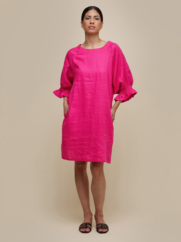 A person stands against a beige background wearing the UCHUU A-LINE DRESS WITH RAGLAN SLEEVES AND ROUND NECK FUSCHIA by UCHUU, a bright pink, loose-fitting A-line dress with three-quarter raglan sleeves and convenient side pockets. The individual is also wearing black, strappy flat sandals and has dark hair pulled back.