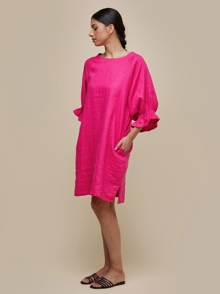 A person stands against a beige background wearing the UCHUU A-LINE DRESS WITH RAGLAN SLEEVES AND ROUND NECK FUSCHIA by UCHUU, a bright pink, loose-fitting A-line dress with three-quarter raglan sleeves and convenient side pockets. The individual is also wearing black, strappy flat sandals and has dark hair pulled back.