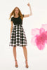 A woman with long hair wears the Signature Silky Knit And Novelty Dot Full Skirt Dress by Joseph Ribkoff, featuring a fit & flare silhouette with knee-length shadow stripes and large polka dots. She stands, one arm raised, against a blurred pink flower background that complements her black high heels.