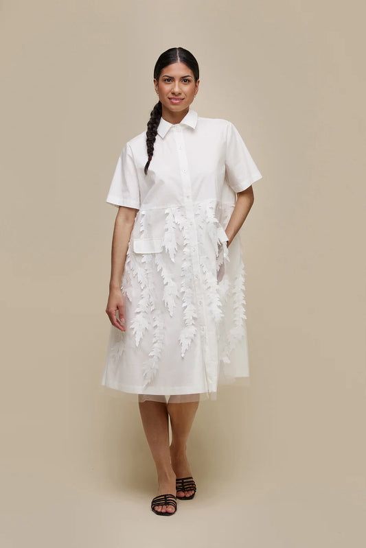 A person with long dark hair in a braid, dressed in a white, short-sleeved, knee-length UCHUU A-LINE BLOUSE STYLE DRESS with leaf-patterned organza appliqué embellishments and invisible side pockets (Product ID: CS24-009), stands against a plain beige background. They wear black slip-on sandals and have a neutral expression on their face.