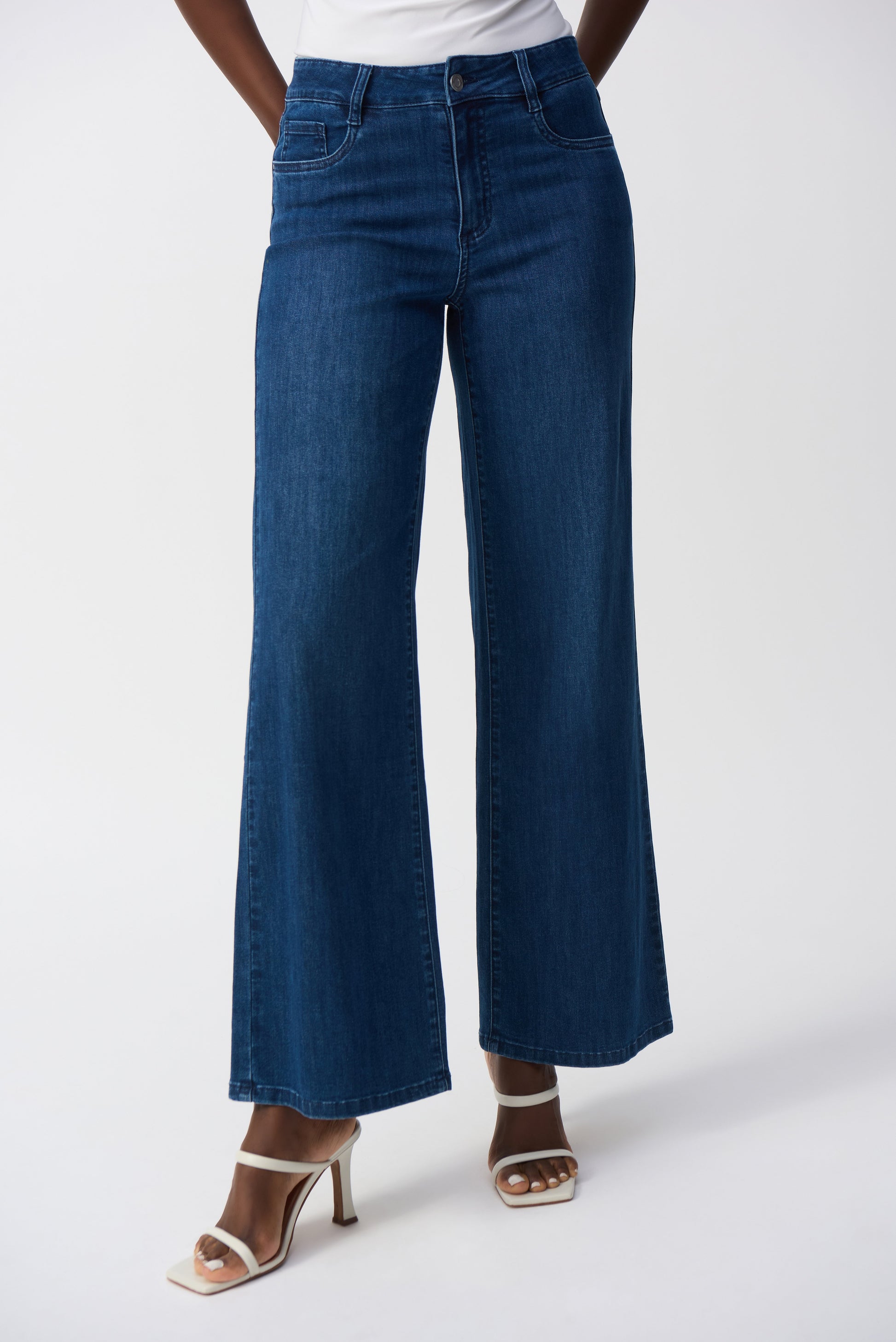 Standing against a plain white background, someone wears Joseph Ribkoff's Goldie Classic Wide-Leg Stretch Jeans in dark blue paired with white open-toe heels.