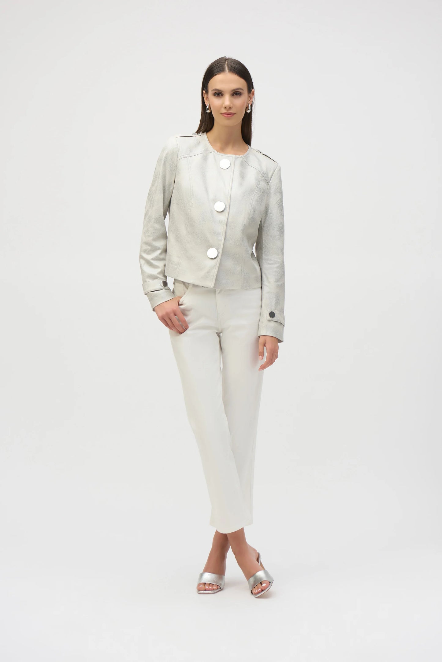 A person with long hair poses against a white background, wearing the Joseph Ribkoff Foiled Faux Suede Boxy Jacket in light gray. The jacket features large metal snap buttons, and they pair it with matching pants and open-toed shoes, resting their hands near their waist.