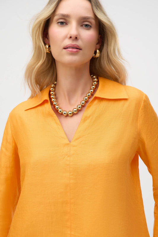 A person with long, wavy blond hair is wearing a bright orange Joseph Ribkoff v-neck blouse and a gold beaded necklace. Complementing the outfit, they have chosen a collarless faux suede jacket and gold hoop earrings against a plain background.