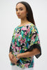 A woman with long dark hair models the Joseph Ribkoff Chiffon Tropical Print Straight Top 252126 and white pants against a plain white background. She complements her look with silver hoop earrings and a bracelet, gazing confidently at the camera.