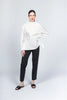 Against a light gray background, a person wears the elegant Italian-crafted Paolo Tricot blouse (Top WT230242 - Pablo Tricot) in O.WHITE with floral embroidery and black pants. Their hair is tied back, black sandals adorn their feet, one hand rests on their hip while the other hangs by their side.