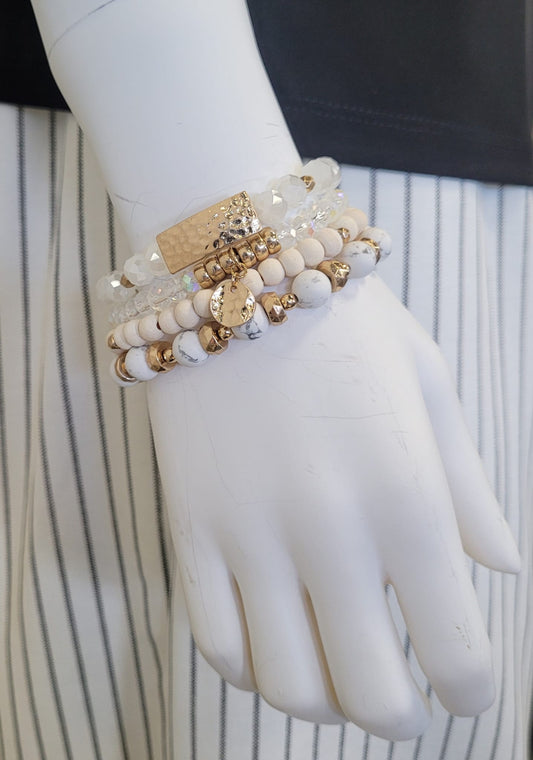 A mannequin hand displays the Gina 26 Bracelet by Elise accessories, a chic piece with layered white, beige, gold, and clear beads, shown with a black top and white skirt with black stripes.