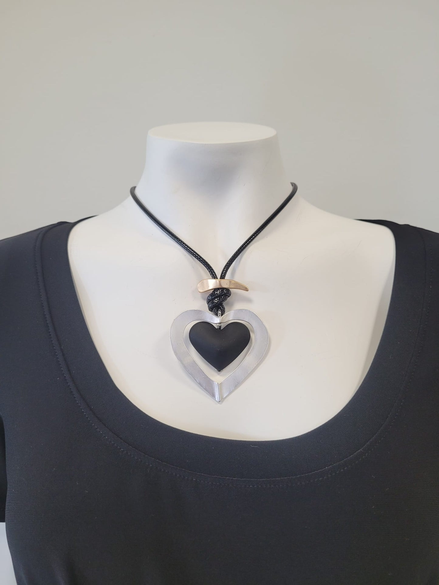 A mannequin in a black scoop-neck top showcases the Sofia 24 Necklace by Elise accessories: a black cord necklace with a large hollow metallic heart pendant and a curved metallic bar accent above it.