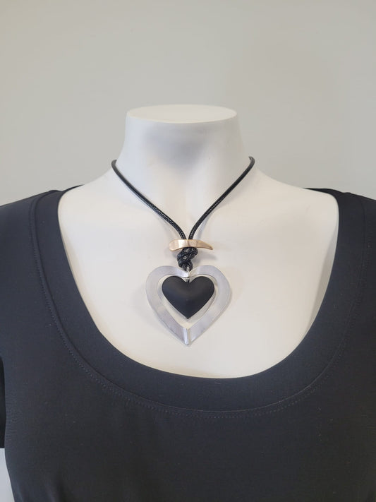 A mannequin in a black scoop-neck top showcases the Sofia 24 Necklace by Elise accessories: a black cord necklace with a large hollow metallic heart pendant and a curved metallic bar accent above it.