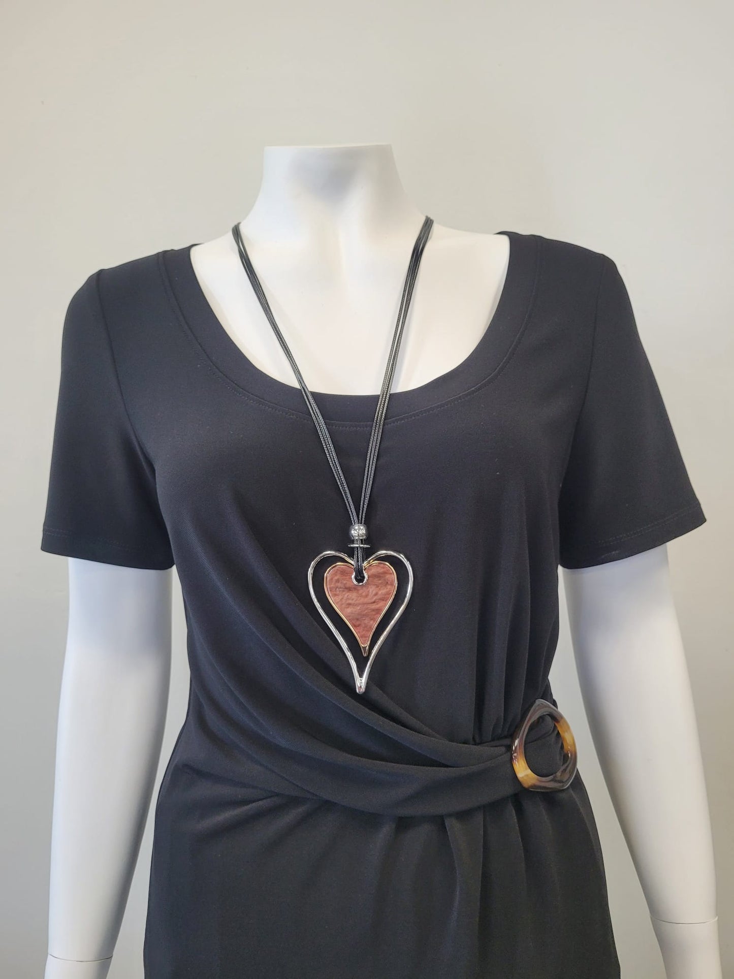 A mannequin in a black short-sleeve top with a side buckle wears the Elise accessories Sofia 13 Necklace, featuring a large pink enamel heart pendant outlined in silver.