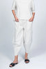 Wearing a stylish white outfit with black vertical stripes, including Pants Striped 33760 by Paolo Tricot and a short-sleeved top, a person casually rests their hands in pockets. Their look is completed by comfy black slide sandals adorned with flowers, all set against a plain light gray backdrop.