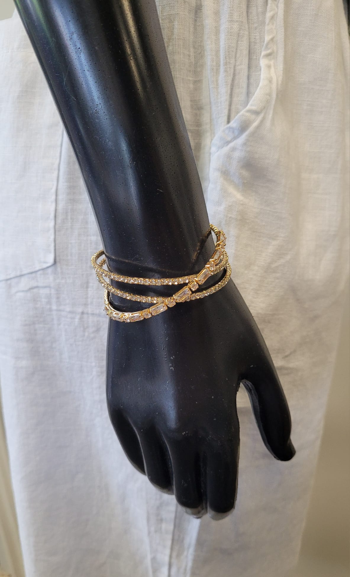 A black mannequin hand displays three gold and crystal bangles, including the Elise accessories Mimosa 13 Bracelet, posed against a backdrop of light linen pants with a visible pocket.