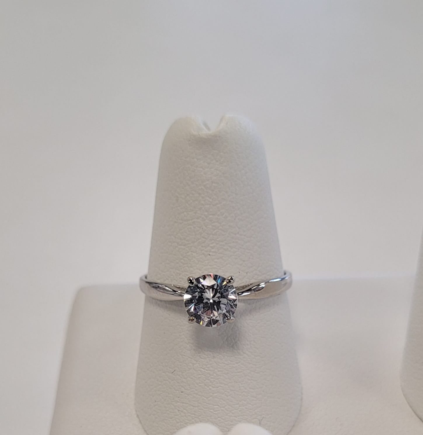 The Elisa 6 Ring by Elise accessories features a single round-cut diamond in a sterling silver prong setting, displayed elegantly on a white textured stand—an ideal accessory for any occasion.