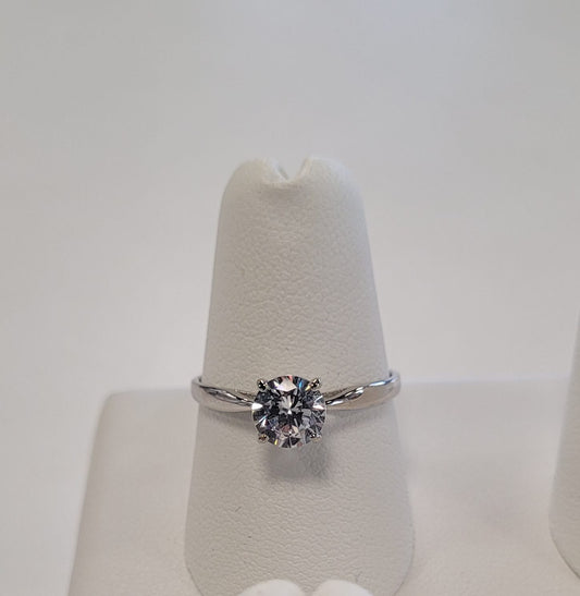 The Elisa 6 Ring by Elise accessories features a single round-cut diamond in a sterling silver prong setting, displayed elegantly on a white textured stand—an ideal accessory for any occasion.