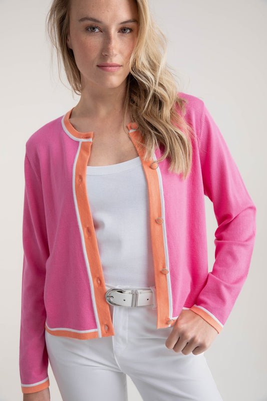 A blonde-haired person wears a MARBLE Sweater 7906 in pink with orange trim over a white shirt, paired with relaxed fit white pants and a matching belt, standing against a neutral background and gazing at the camera.