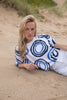 On a sandy beach, a person with long blonde hair relaxes, dressed in MARBLE's Sweater Top 7809 MARBLE and white pants, creating a luxurious vibe. The scene is set against grass and sparse vegetation.