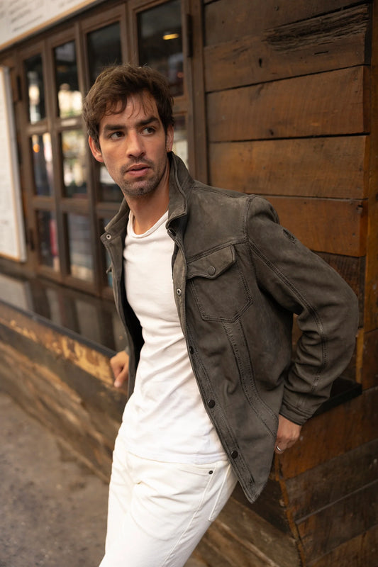 A man with tousled brown hair wears the MAURITIUS LEATHER Sedro RF Leather Jacket in Khaki over a white t-shirt and white pants, leaning against a wooden wall and gazing thoughtfully to the side.