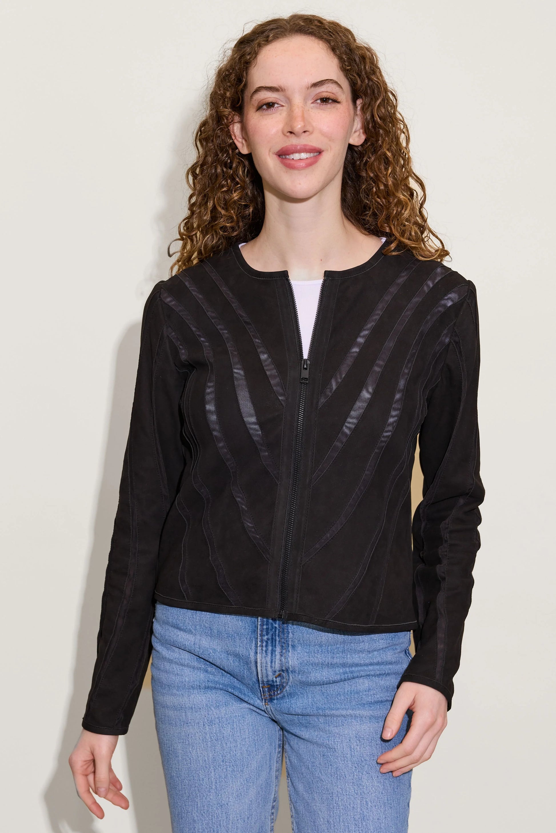 A woman with curly brown hair wears the MAURITIUS LEATHER Mira RF Leather Jacket—featuring subtle shiny stripes and a breathable mesh lining—over a white shirt and blue jeans, smiling at the camera against a plain light background.