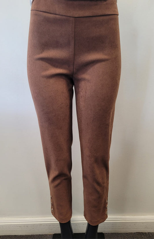 A person wearing Bali Corp's Classic Brown Pant—high-waisted, slim-fit ankle pants in polyester-spandex with button accents near the hem—stands against a plain white wall.