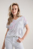 A person with long blonde hair wears the Top T-shirt 7848 MARBLE by MARBLE, featuring a light, relaxed fit and circular perforated pattern, paired with white pants — ideal for everyday wear. They stand against a plain background, gazing at the camera with a neutral expression.