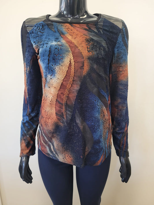 A mannequin wears the Bali Corp Top 8693, featuring mesh shoulders and long sleeves with an abstract wavy pattern in blue, orange, brown, and black, paired with dark pants.