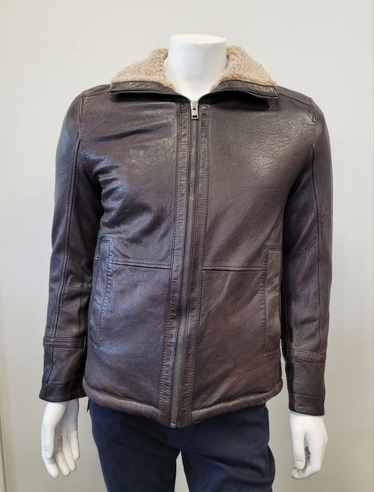 A mannequin wears a MAURITIUS LEATHER Nuuko UF Leather Jacket in dark brown with a light shearling collar, front zipper, and two large pockets, paired with dark blue pants, posed against a plain light background.