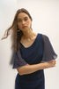 A woman poses against a white wall in the Signature Silky Knit and Chiffon Sheath Dress by Joseph Ribkoff. With butterfly sleeves and an asymmetrical neckline, she wears hoop earrings and gazes softly, her long hair gently moved by a breeze.