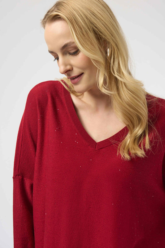 A woman with long blonde hair smiles gently while wearing the Joseph Ribkoff Sequined Sweater Knit Boxy Top 244921F25, a loose-fitting red top with a V-neckline, against a plain light background.