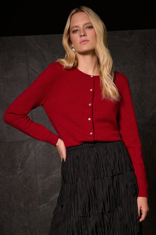 A woman with long blonde hair models the Joseph Ribkoff Cashmere Button-Front Crew Neck Cardigan 254951 in red, paired with a high-waisted black pleated skirt, against a dark textured backdrop.