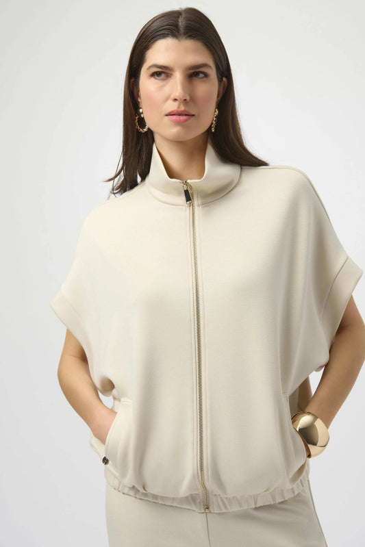 A woman with long dark hair models the Joseph Ribkoff Scuba Jersey Boxy Jacket 254145 in cream, featuring short sleeves and a stand collar. She accessorizes with gold hoops and bracelet, posing hands-in-pocket against a light background.